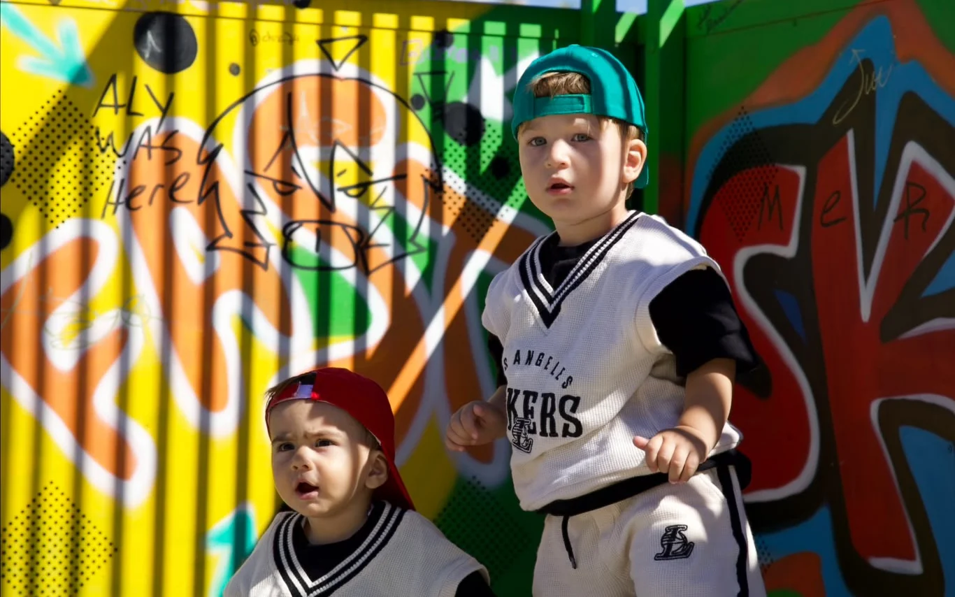 Gueto Kids takım giyen iki çocuk, kaykaylarıyla grafitili bir duvarın önünde